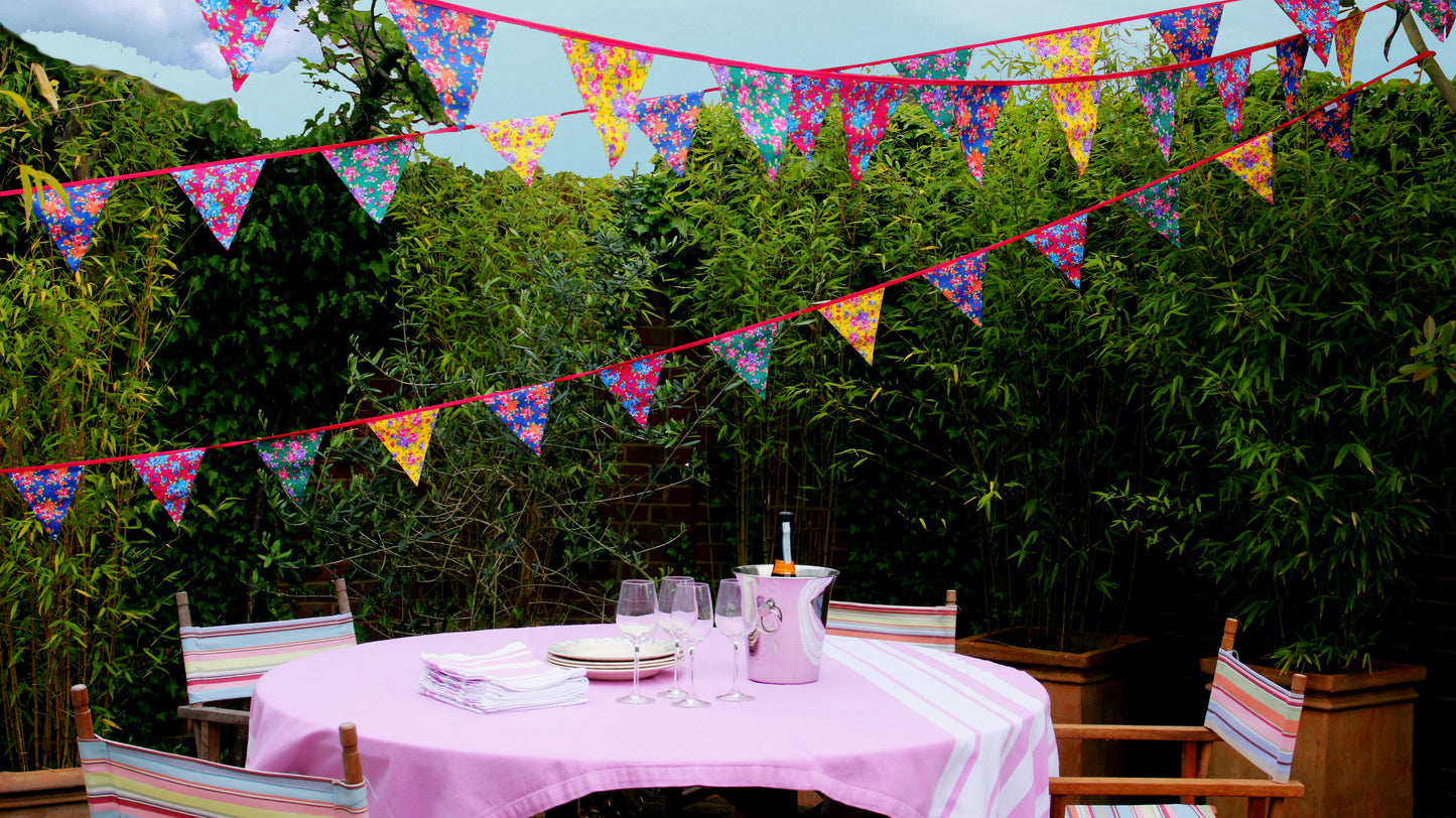 Outdoor setting with a table and chairs under colourful floral bunting against a green forest background