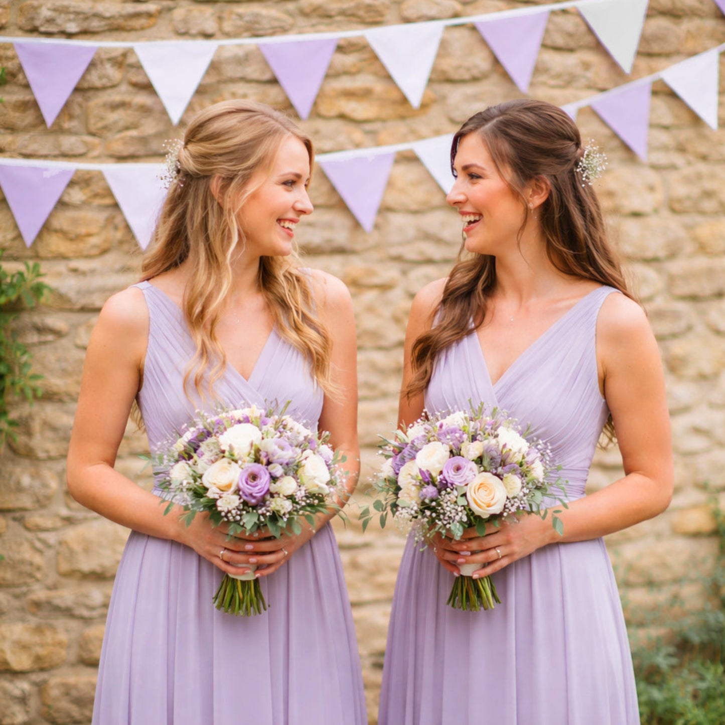 Two women in lavender dresses holding floral bouquets in front of a stone wall with a purple and white banner.