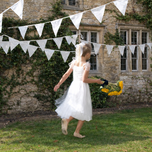 Young girl in a white dress holding flowers under a string of white flags in front of a stone building.